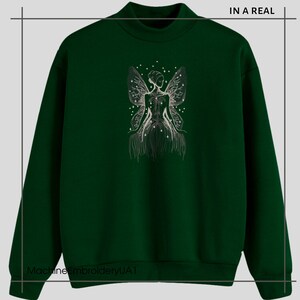 May include: A dark green sweatshirt with a white embroidered design of a fairy with wings and stars. The text "IN A REAL" is in the top right corner of the image.
