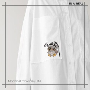 Op de afbeelding: Een wit overhemd met knopen en een zak aan de linkerkant. De zak heeft een machine-geborduurd ontwerp met een grijze uil die een gestreepte slaapmuts draagt. De tekst "IN A REAL" is gedrukt in de rechterbovenhoek van het overhemd. De tekst "MachineEmbroideryUA1" is gedrukt in de linkerbenedenhoek van het overhemd.