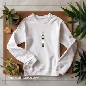 May include: White crewneck sweatshirt with an embroidered nutcracker design in light blue, gold, and black. The sweatshirt features the text "MachineEmbroideryUA1" at the bottom. The design is centered on the chest.
