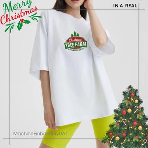 K&ouml;nnte beinhalten: Wei&szlig;es Oversize-T-Shirt mit einem gr&uuml;n-roten Weihnachtsbaum-Farm-Logo. Das Logo lautet "Christmas Tree Farm Cut and Carry". Das Model tr&auml;gt das Shirt mit gelber Shorts.