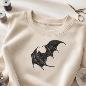 May include: A beige crewneck sweatshirt featuring a detailed black embroidered bat design on the chest. The bat is depicted with spread wings and visible white fangs. The garment has a ribbed collar and long sleeves, presented with sewing supplies.