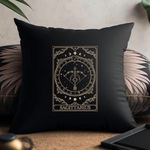 Pu&ograve; includere: Cuscino nero con un disegno ricamato in oro di un simbolo zodiacale, un arco e una freccia, e la parola "SAGITTARIO".
