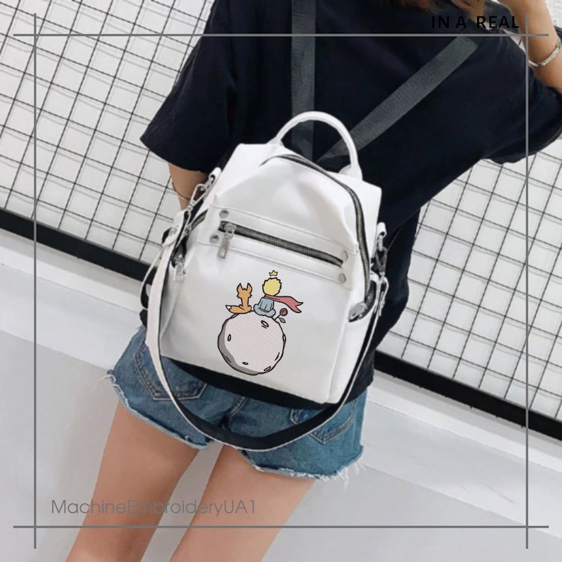 May include: White faux leather backpack with a zippered pocket and a graphic of The Little Prince sitting on an asteroid with a fox. The backpack has a black adjustable strap.