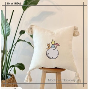 May include: A white pillow with a beige tassel on each corner. The pillow features an embroidered image of The Little Prince sitting on an asteroid with a fox.