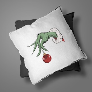 May include: A white pillow with a green hand holding a red Christmas ornament. The hand is wearing a white fur cuff. The pillow is on a black pillow.