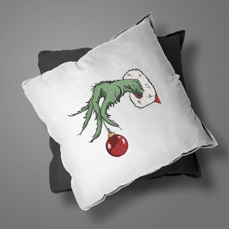 May include: A white pillow with a green hand holding a red Christmas ornament. The hand is wearing a white fur cuff. The pillow is on a black pillow.