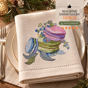 May include: A beige linen napkin featuring a machine embroidery design of stacked colorful macarons in shades of pink, green, and blue, accented with small floral sprigs and leaves. The text reads MACHINE EMBROIDERY DESIGN and DIGITAL DOWNLOAD.