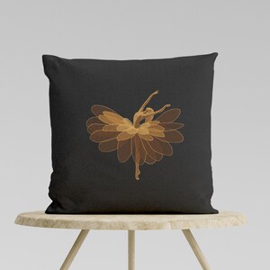 May include: Black square throw pillow featuring an embroidered design of a ballerina. The ballerina is depicted with a flower-like skirt and outstretched arms, in shades of gold. The pillow is resting on a light-colored wooden table.