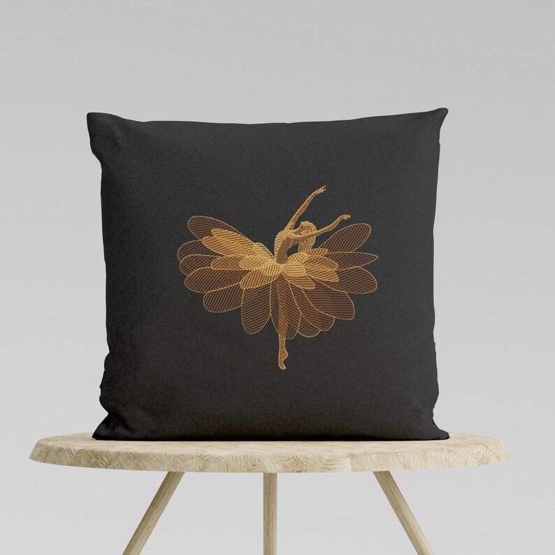 May include: Black square throw pillow featuring an embroidered design of a ballerina. The ballerina is depicted with a flower-like skirt and outstretched arms, in shades of gold. The pillow is resting on a light-colored wooden table.