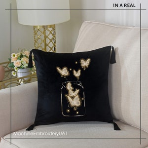 May include: Black velvet pillow with a white embroidered design of butterflies flying out of a glass jar. The pillow has tassels on the corners. "MachineEmbroideryUA1" is written in white at the bottom of the pillow.