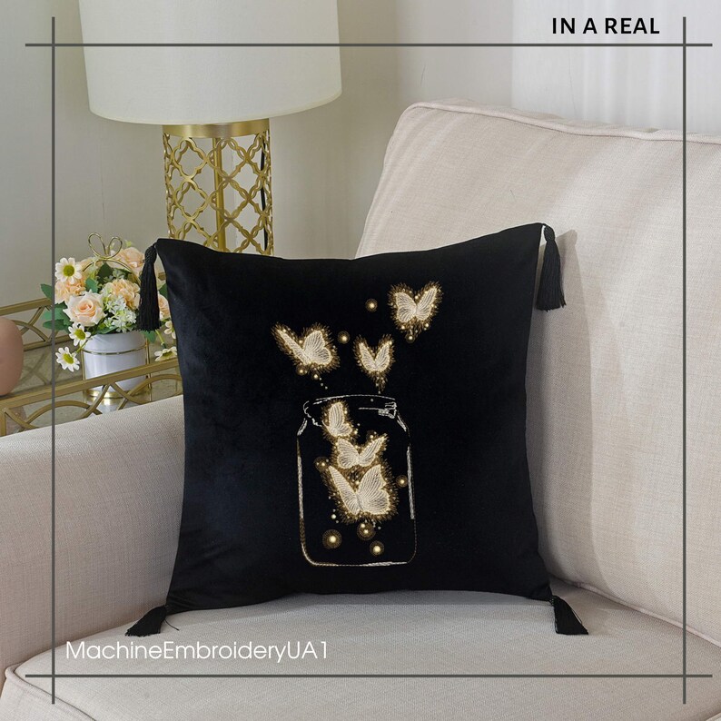 May include: Black velvet pillow with a white embroidered design of butterflies flying out of a glass jar. The pillow has tassels on the corners. "MachineEmbroideryUA1" is written in white at the bottom of the pillow.