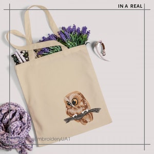 May include: Cream-coloured tote bag with an embroidered owl design. The owl is brown and tan, with yellow eyes, perched on a branch. The bag contains lavender and a patterned item. Text on the bag reads "IN A REAL".