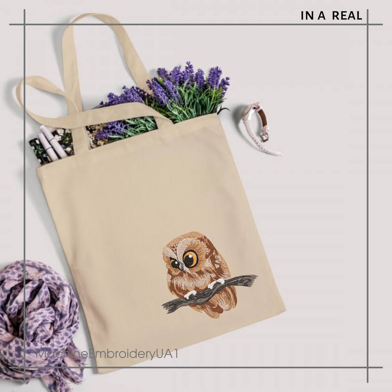 May include: Cream-coloured tote bag with an embroidered owl design. The owl is brown and tan, with yellow eyes, perched on a branch. The bag contains lavender and a patterned item. Text on the bag reads "IN A REAL".