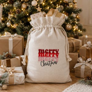 May include: A beige canvas drawstring gift sack featuring the embroidered text merry merry merry Christmas in stacked red and pink wavy lettering, with a cinched top and thick rope ties, positioned in front of a decorated Christmas tree.