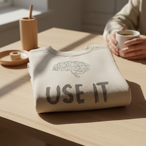 Może przedstawiać: Złożona beżowa bluza z nadrukiem m&oacute;zgu i napisem "USE IT" w ciemnoszarym kolorze. Bluza leży na jasnym drewnianym stole. Na stole znajdują się r&oacute;wnież drewniany pojemnik na oł&oacute;wki i mały talerz.
