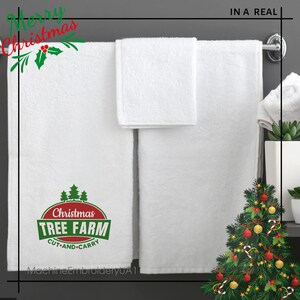 K&ouml;nnte beinhalten: Wei&szlig;e Badet&uuml;cher mit einem gestickten Weihnachtsbaum-Farm-Logo in Rot und Gr&uuml;n auf einem Handtuch. Die Handt&uuml;cher h&auml;ngen an einem Handtuchhalter mit einem kleinen Weihnachtsbaum im Hintergrund.