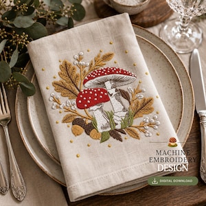 Pode incluir: Um guardanapo de linho bege com bordado à máquina de dois cogumelos vermelhos e brancos rodeados por folhas de carvalho castanhas, bolotas e pequenas bagas brancas. O texto "MACHINE EMBROIDERY DESIGN" e "DIGITAL DOWNLOAD" está impresso no canto inferior direito.