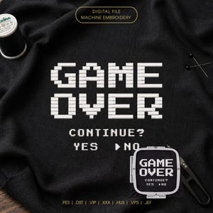 Może przedstawiać: Cyfrowy plik do haftu maszynowego z białym, pikselowym napisem GAME OVER, CONTINUE? YES NO na czarnej tkaninie. Projekt zawiera mały tamborek z tym samym tekstem, szpulkę czarnej nici oraz metalowe nożyczki.