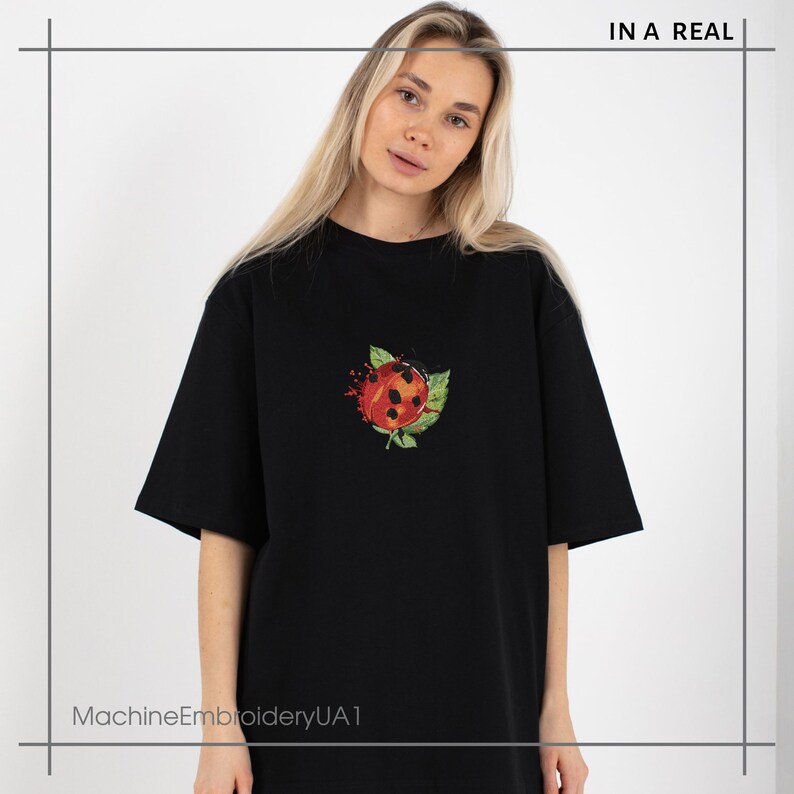 Może przedstawiać: Czarny T-shirt z haftowaną czerwoną i czarną biedronką z przodu. Biedronka jest otoczona zielonymi liśćmi i ma za sobą rozprysk pomarańczowej farby. Tekst "IN A REAL" znajduje się w prawym g&oacute;rnym rogu obrazu. Tekst "MachineEmbroideryUA1" znajduje się w lewym dolnym rogu obrazu.