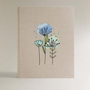 May include: A beige linen-textured journal with embroidered floral design. The embroidery features blue and green stylized flowers with black stems and accents. The journal has a clean, minimalist aesthetic.