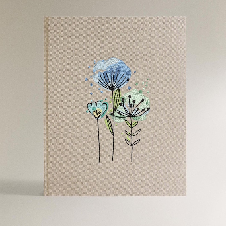May include: A beige linen-textured journal with embroidered floral design. The embroidery features blue and green stylized flowers with black stems and accents. The journal has a clean, minimalist aesthetic.