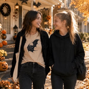 May include: A beige t-shirt featuring a black bat graphic, worn under a black button-down shirt with black jeans and a beige tote bag, alongside a black hooded sweatshirt with a black backpack, both styled for a casual autumn aesthetic.