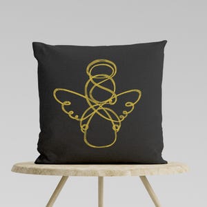 May include: A black cushion with a gold embroidered angel design. The angel is depicted in a simple, minimalist style with a halo and wings.