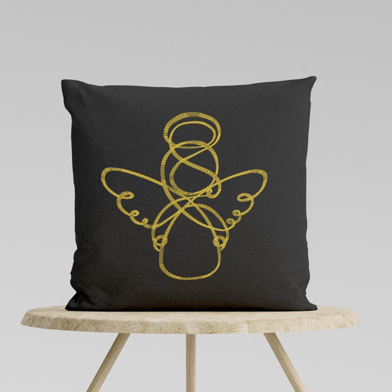 May include: A black cushion with a gold embroidered angel design. The angel is depicted in a simple, minimalist style with a halo and wings.