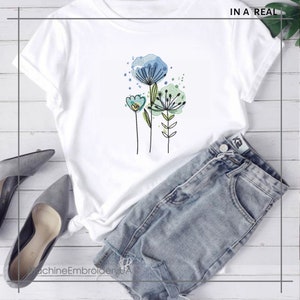 May include: White t-shirt featuring an embroidered floral design in shades of blue, green, and yellow. The shirt is paired with denim shorts and gray heels. The text "IN A REAL" is visible in the upper right corner.