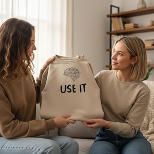 Może przedstawiać: Beżowa bluza z czarną grafiką m&oacute;zgu i napisem "USE IT". Bluza jest trzymana przez dwie osoby. Tło obejmuje sofę, regał i okno.