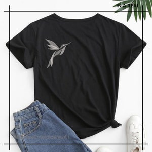 Puede incluir: Camiseta negra de manga corta con un colibr&iacute; plateado bordado en el lado izquierdo del pecho. La camiseta est&aacute; sobre un par de pantalones vaqueros azules y zapatillas blancas.