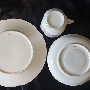 May include: A white teacup and saucer set with a pink floral design. The bottom of the saucer and cup have the number "1974" printed in red.