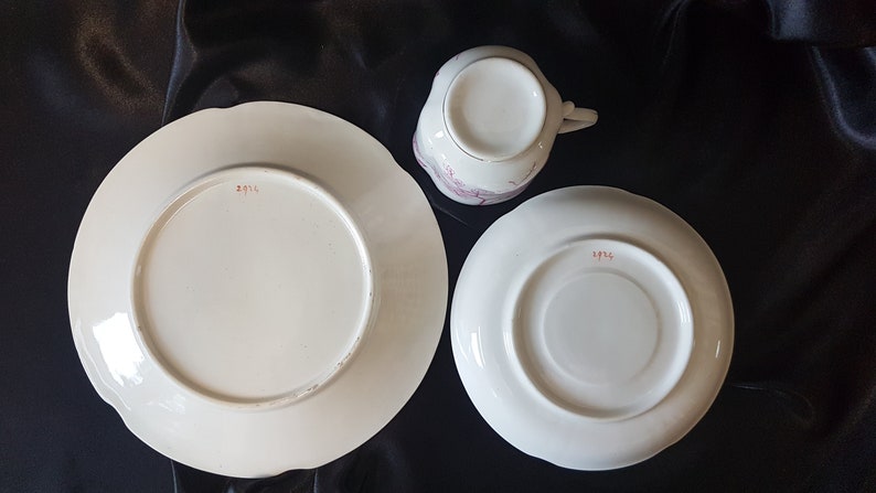 May include: A white teacup and saucer set with a pink floral design. The bottom of the saucer and cup have the number "1974" printed in red.