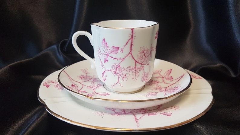 May include: A white teacup and saucer set with a pink floral design. The cup and saucer have a gold trim around the edge.