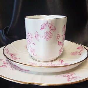 May include: A white teacup and saucer set with a pink floral pattern and gold trim. The set is on a black background.