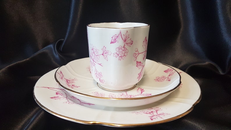 May include: A white teacup and saucer set with a pink floral pattern and gold trim. The set is on a black background.
