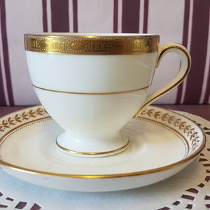 May include: A white teacup and saucer with gold trim. The cup has a delicate floral design and the saucer has a simple gold band.