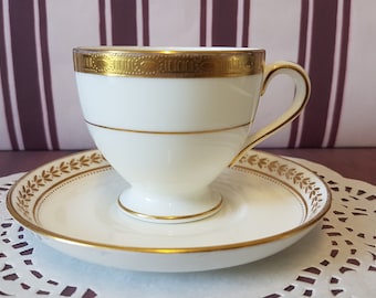 Puede incluir: Una taza de té y un platillo blancos con ribetes dorados. La taza tiene un delicado diseño floral y el platillo tiene una simple banda dorada.