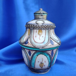 May include: A ceramic jar with a silver lid and intricate painted designs in shades of blue, green, white, and yellow. The jar has a round body and a narrow neck.