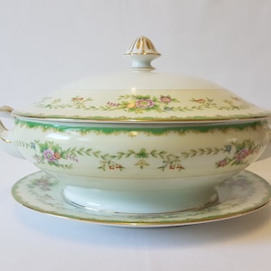 Może przedstawiać: Biała porcelanowa miska do serwowania z pokrywką, ozdobiona kwiatowym wzorem w odcieniach zieleni, różu i żółci. Miska ma złotą obwódkę i pasującą podstawę.