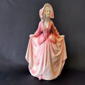 Może przedstawiać: Porcelanowa figurka kobiety w różowej sukni i kapeluszu. Figurka ma długą, powłóczystą spódnicę i dopasowany gorset. Kobieta trzyma spódnicę obiema rękami. Kapelusz ma szerokie rondo. Figurka stoi na ciemnym tle.