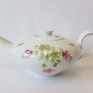 May include: A white porcelain teapot with a floral design featuring pink flowers and green leaves. The teapot has a lid and a handle.