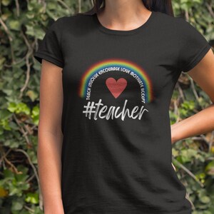 May include: Black t-shirt with a rainbow arc and a red heart. The arc has the words "Teach Inspire Encourage Love Motivate Accept" written on it. The text "#teacher" is below the heart.