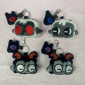 May include: Four plush keychains featuring cartoon characters with different expressions. Each keychain has a black and white character with a different coloured eye and a black and white cat or bat hanging from the top of the keychain.