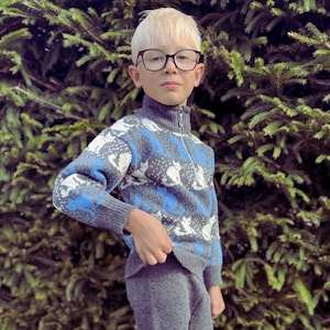 May include: A child in a grey knitted sweater and trousers set. The sweater has a pattern of white cats and blue details. The child is also wearing glasses and grey and white trainers.