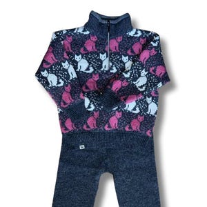 Op de afbeelding: Een tweedelige kinderoutfit. De top is een donkergrijze trui met een hoge kraag en een korte ritssluiting. De trui heeft een patroon van roze en witte katten. De bijpassende broek is effen donkergrijs.