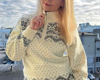 Pode incluir: Camisola creme com gola alta e fecho de correr a meio. A camisola apresenta um padrão de flocos de neve cinzentos e desenhos geométricos. As mangas e o corpo são pontilhados com pequenas formas cinzentas. A pessoa está a usar calças de ganga azuis.