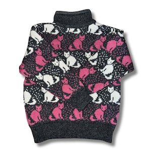 May include: A grey turtleneck jumper featuring a repeating pattern of white and pink cats. The jumper has a dark grey collar, cuffs, and hem. The cat pattern is set against a black background with white dots.