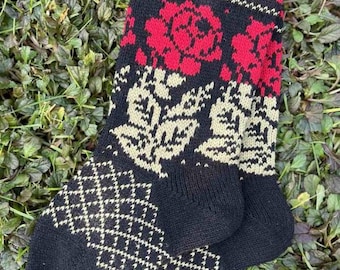 May include: A pair of black knit socks with a floral pattern in red and off-white. The socks feature a diamond pattern on the foot and a wavy design at the top. The socks are lying on a bed of green leaves.