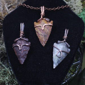 May include: Three arrowhead pendants on a black display stand. The central pendant is a light brown color, while the other two are a darker brown and a light grey. Each arrowhead is wrapped in copper wire and suspended from a copper chain.